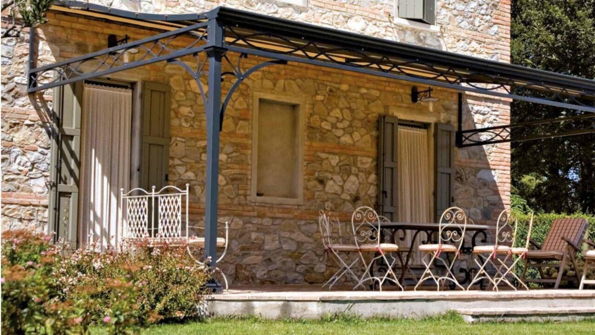 A stone house with green shutters features a metal pergola covering a patio with a white bench and a dining set of chairs and a table, surrounded by greenery and lawn.