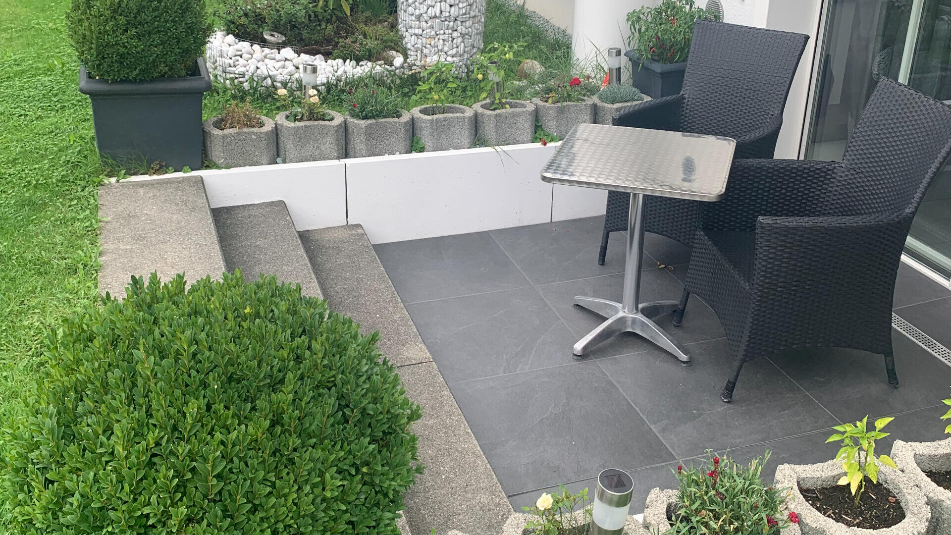 Eine kleine Veranda mit zwei schwarzen Korbstühlen und einem Metalltisch steht neben einer Steintreppe. Drumherum stehen Topfpflanzen, ein grüner Busch und ein Ziergarten mit weißen Steinen und Blumen.