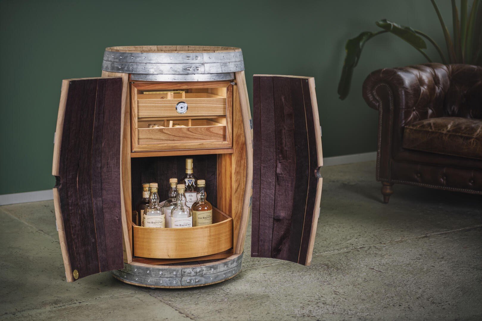 Ein offenes Holzfass, das als Schnapsschrank umfunktioniert wurde, mit zwei Regalen für Flaschen und Gläser, steht auf einem Betonboden neben einem braunen Ledersofa und einer grünen Wand.