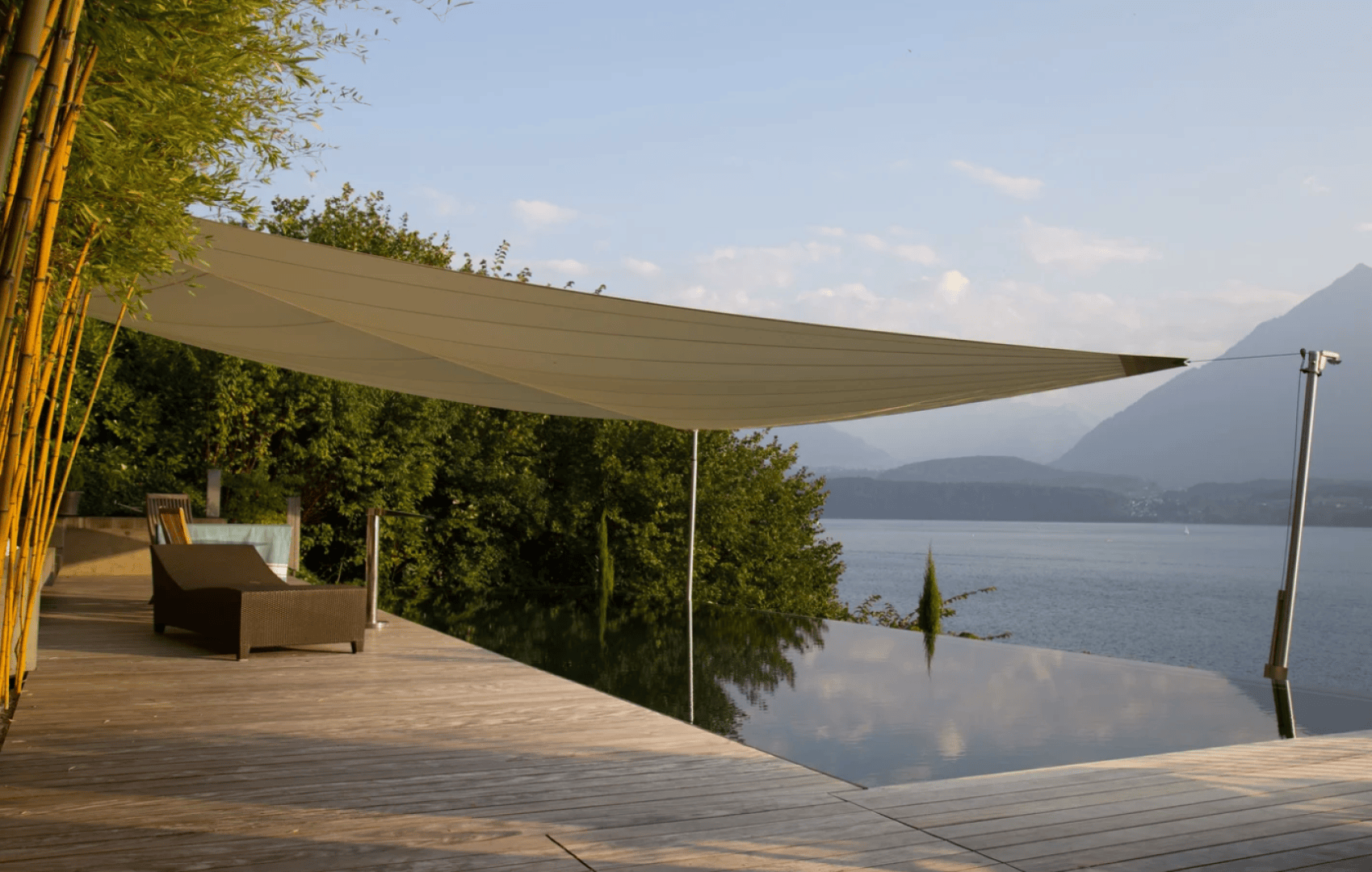 A modern wooden deck with lounge chairs overlooks an infinity pool and a large lake, surrounded by greenery and mountains. A white shade sail provides cover above part of the deck.
