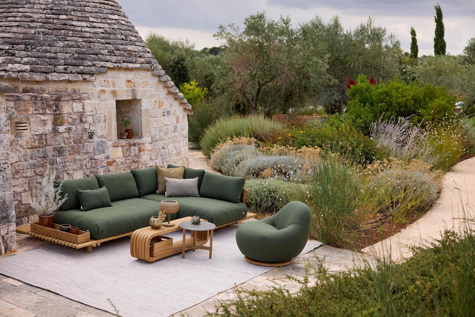 An outdoor seating area with a green upholstered sofa and armchair, a small coffee table and a rug is located next to a rustic stone house, surrounded by lush greenery and garden paths.