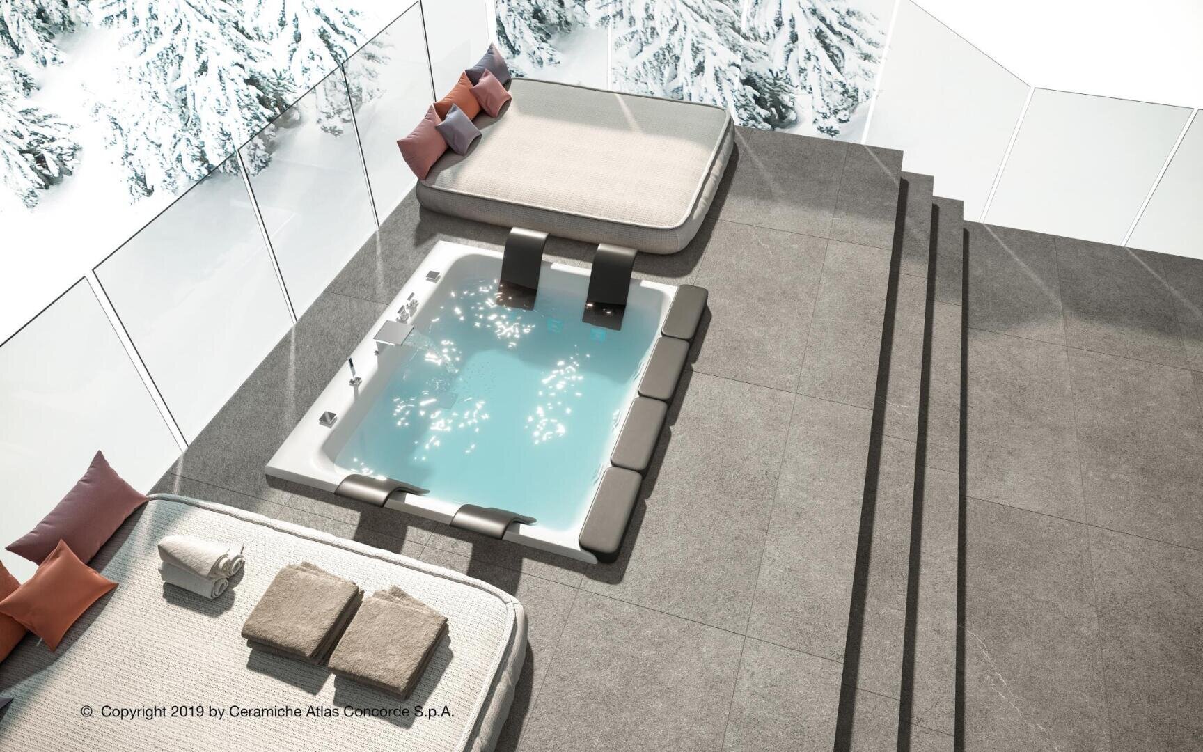 A modern, elevated outdoor hot tub with built-in seats is surrounded by large gray tiles. Nearby are two lounge beds with cushions and pillows. Through glass panels, snowy mountains are visible in the background.