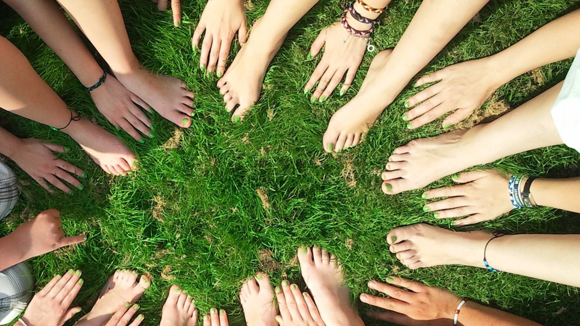 Eine Gruppe von Menschen, die im Kreis auf einer Wiese sitzen und ihre nackten Füße und Hände in die Mitte strecken, mit bemalten Nägeln und verschiedenen Armbändern.