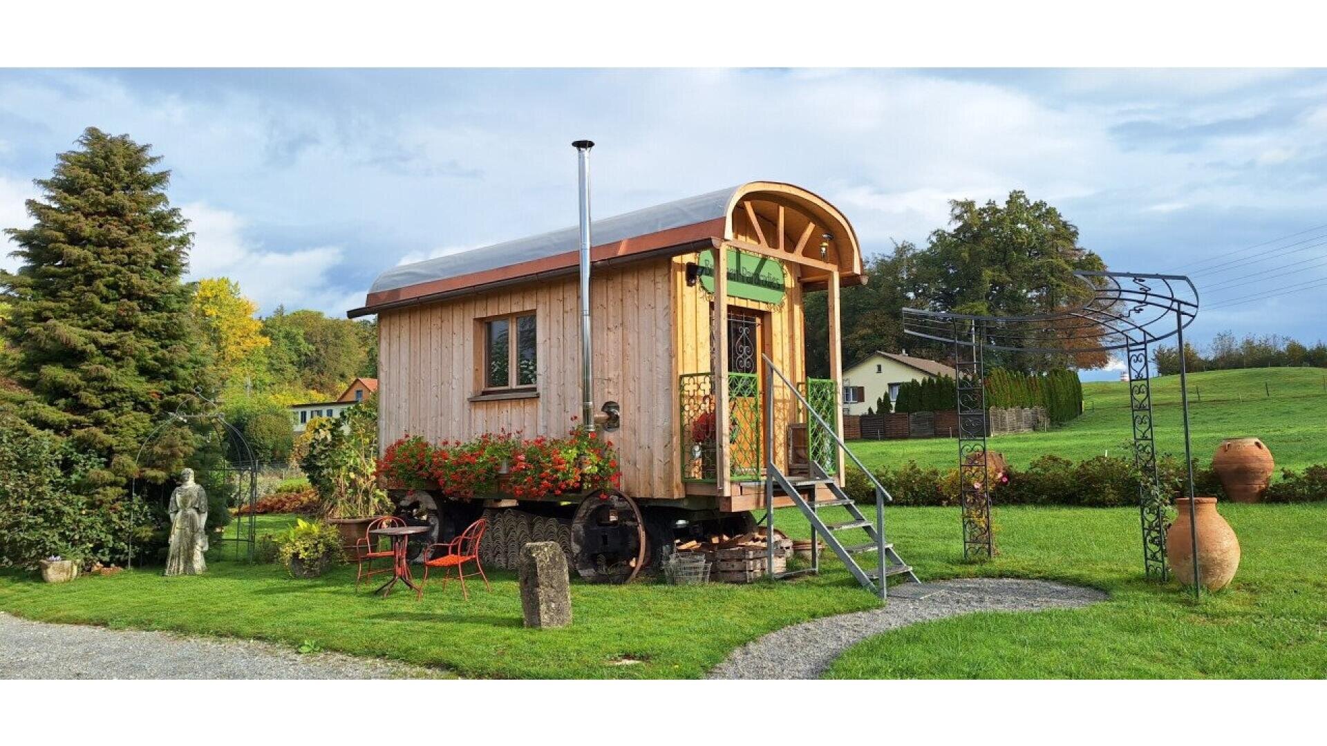 A small wooden tiny house with a curved roof sits on wheels in a grassy yard, decorated with red flowers and outdoor furniture. A metal staircase leads to the door, and trees and pottery jars surround the area.