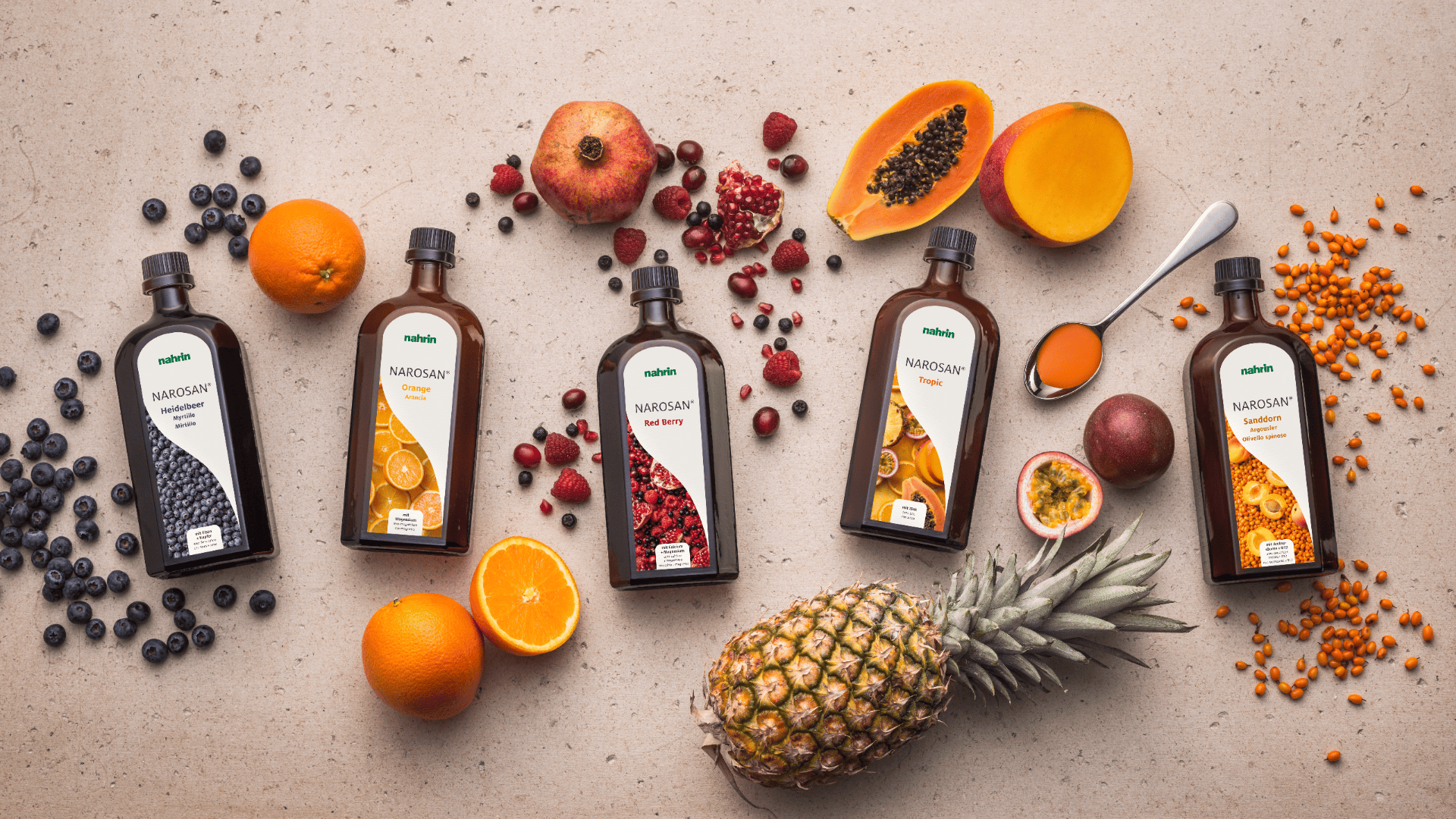 Five brown bottles of fruit concentrate stand on a light-colored surface, surrounded by fresh fruit such as oranges, pineapple, blueberries, papaya, pomegranates and berries, with a spoonful of orange liquid next to them.