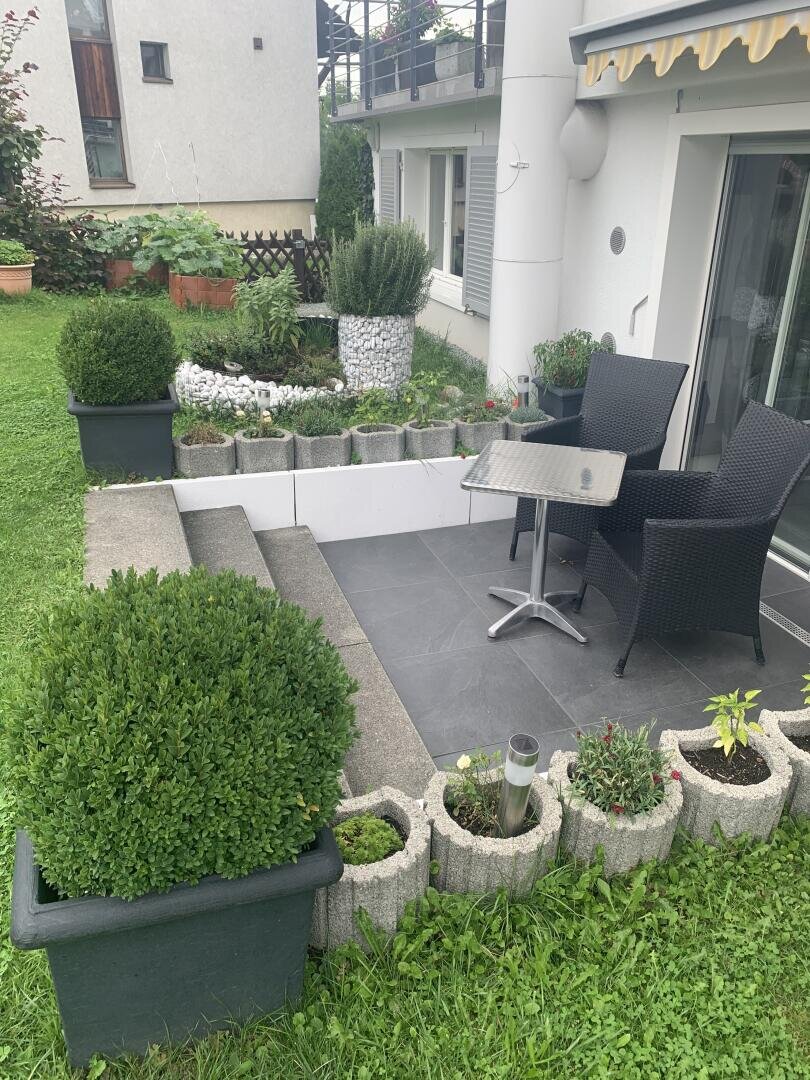 Eine kleine Veranda mit zwei schwarzen Korbstühlen und einem Tisch, umgeben von Topfpflanzen und Grünzeug. Betontreppen führen hinunter zu einer Rasenfläche in einem gepflegten Garten neben einem modernen weißen Gebäude.