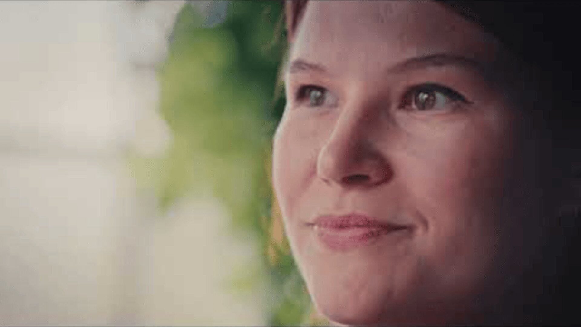 A woman with fair skin and brown hair smiles softly, looking slightly upward. The background is blurred with hints of greenery and sunlight.