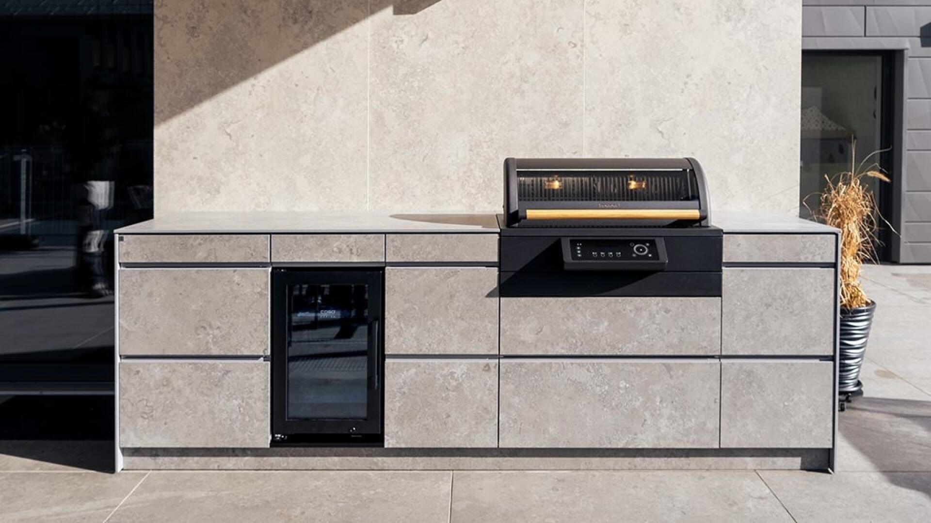 Eine moderne Außenküche mit Schränken aus hellem Stein, einem eingebauten Minikühlschrank mit Glastür und einem eleganten Grill mit offenem Deckel und zwei Lampen im Inneren, der auf einer Steinterrasse steht.