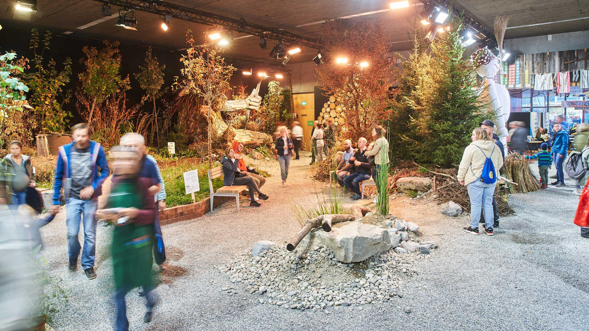 Die Besucher gehen und sitzen in einem überdachten Garten mit Bäumen, Steinen, Pflanzen und natürlichen Dekorationen unter hellen Lichtern. Es herrscht eine lebhafte Atmosphäre, in der mehrere Besucher die Ausstellung erkunden.
