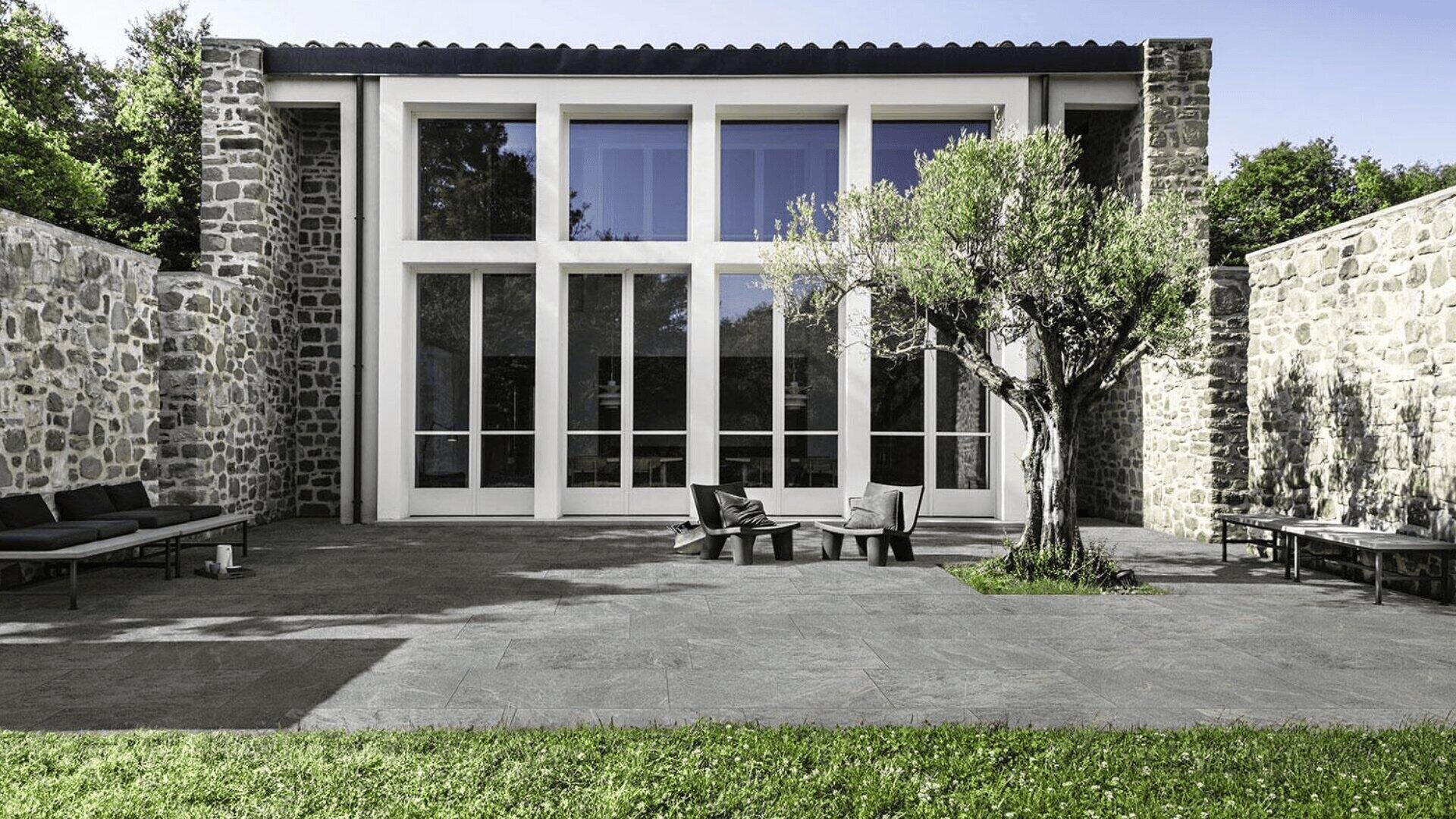 Modern stone house with large windows and a minimalist patio featuring outdoor chairs, a tree in the center, and surrounding stone walls. Green grass borders the stone-paved yard.