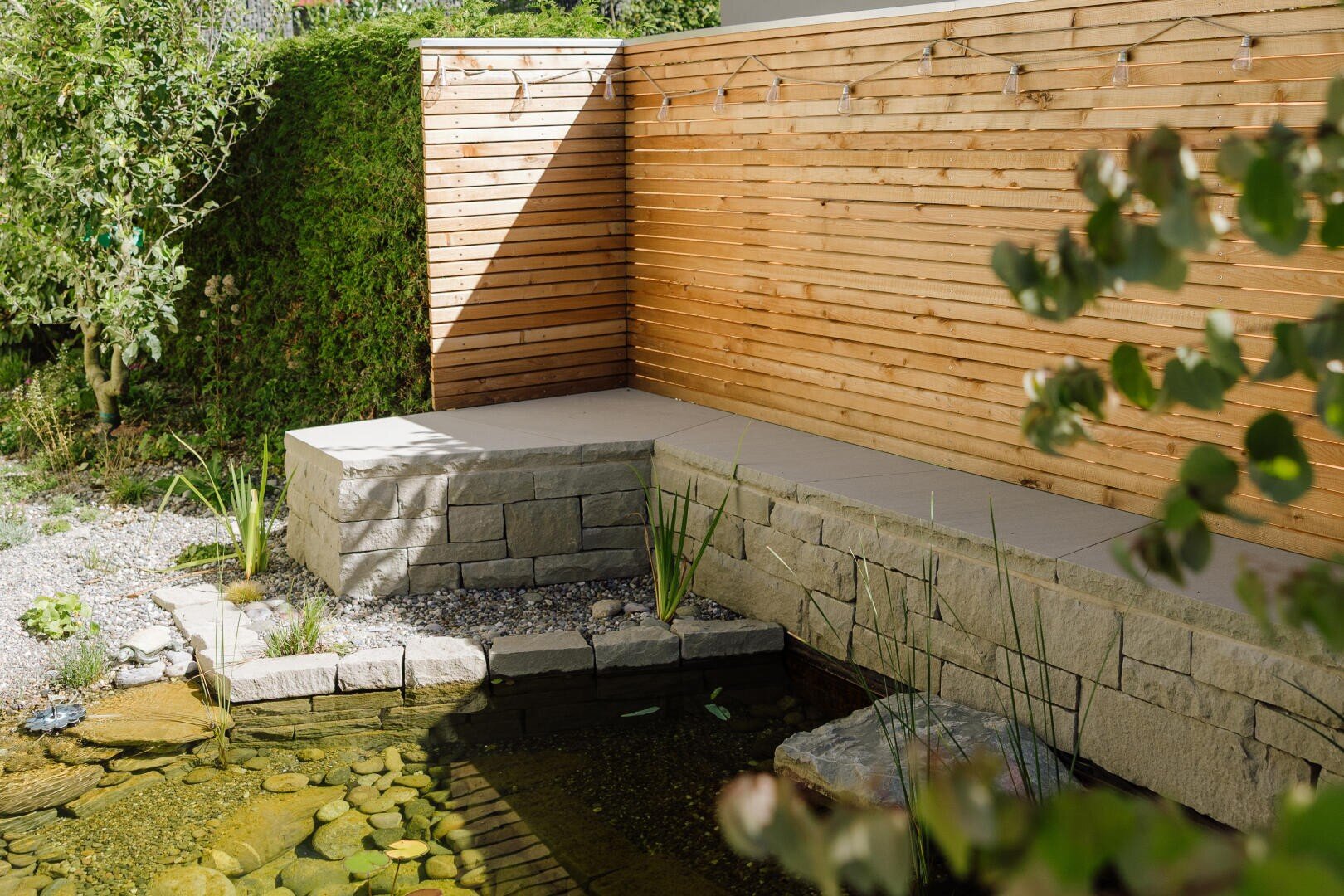 Ein kleiner Gartenteich mit klarem Wasser und Steinrändern liegt neben einer Eckbank aus Beton und Stein, hinter der ein horizontaler Holzzaun mit Lichterketten angebracht ist. Grünpflanzen und Sträucher umgeben den Bereich.