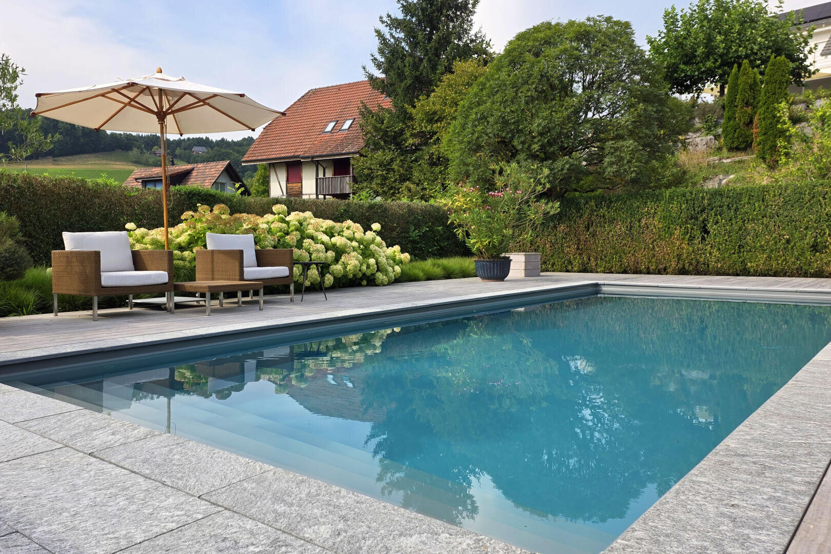 Ein moderner Pool im Hinterhof, umgeben von Steinfliesen, mit zwei Korbstühlen und einem Sonnenschirm auf einer Holzterrasse. Im Hintergrund sind üppige grüne Büsche, Bäume und ein Haus mit rotem Dach zu sehen.