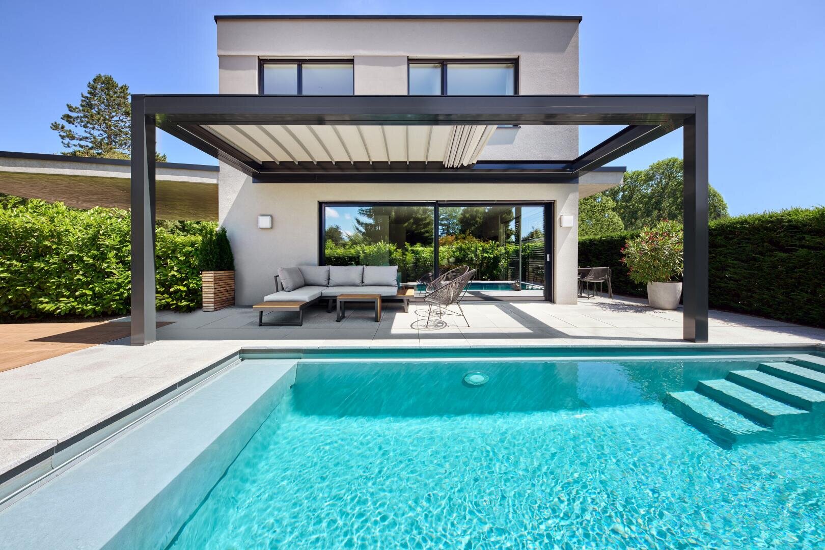 Modernes Haus mit einer Pergola-überdachten Terrasse, Sitzgelegenheiten im Freien und einem klaren blauen Swimmingpool im Vordergrund, umgeben von Grün und Bäumen unter einem strahlenden, sonnigen Himmel.