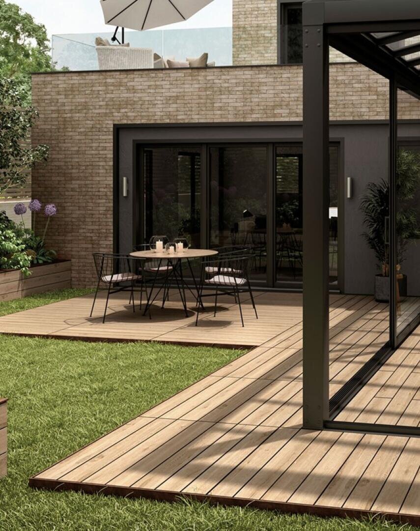 Ein moderner Hinterhof mit einer Holzterrasse, einem Esstisch und Stühlen im Freien sowie Glastüren, die in ein Haus mit Backsteinmauern und großen Fenstern führen. Grünes Gras und Pflanzen umrahmen den Innenhof.