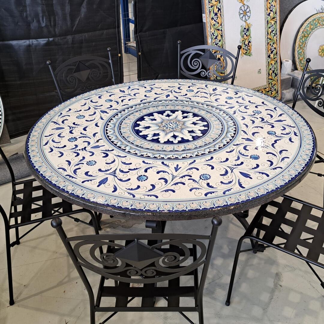 A round table with a detailed blue and white mosaic pattern is surrounded by four black wrought-iron chairs with decorative backrests and seats, set in an interior space on a light-colored floor.