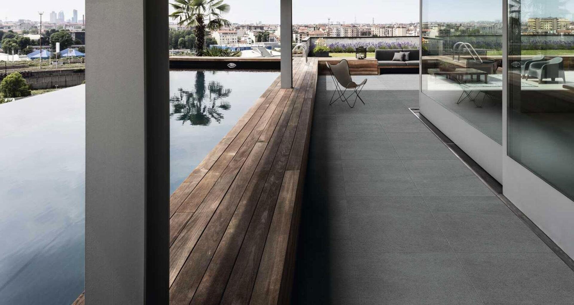Modern terrace with a wooden bench beside a rectangular pool, glass wall on the right, a lounge chair, and cityscape in the background under a partly cloudy sky.
