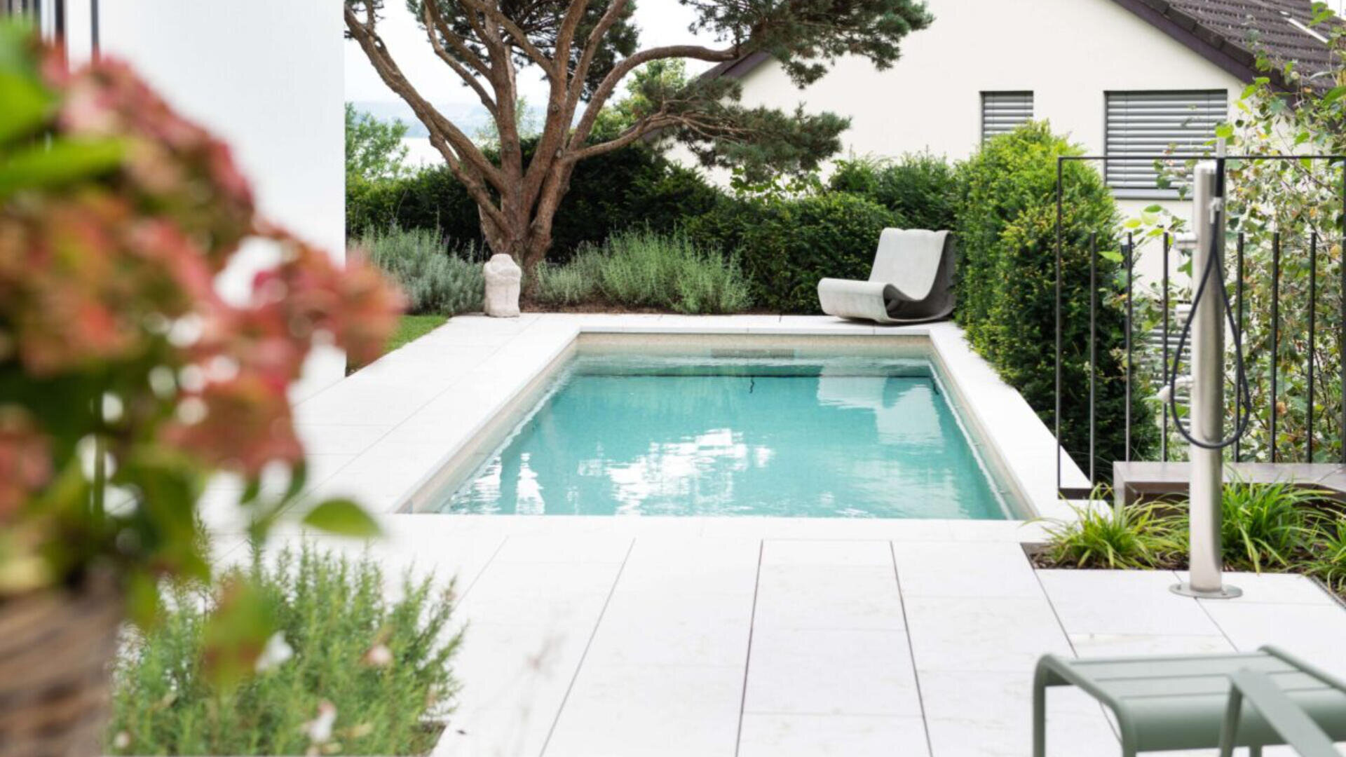 Ein kleiner, rechteckiger Gartenpool mit klarem Wasser ist von weißen Fliesen und üppigem Grün umgeben. Ein einzelner Liegestuhl steht am Ende des Pools, neben einem Baum und einigen Sträuchern.