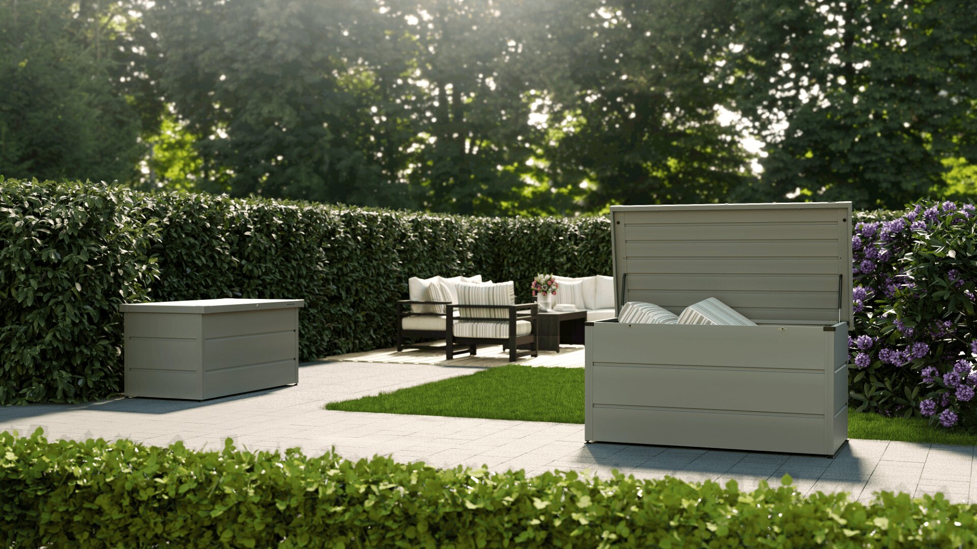 Auf der gepflegten Terrasse befinden sich zwei hellgraue Aufbewahrungsboxen, von denen eine offen ist und Kissen enthält, sowie ein Sitzbereich mit weißen gepolsterten Stühlen und einem Tisch, umgeben von üppigem Grün und blühenden Büschen.