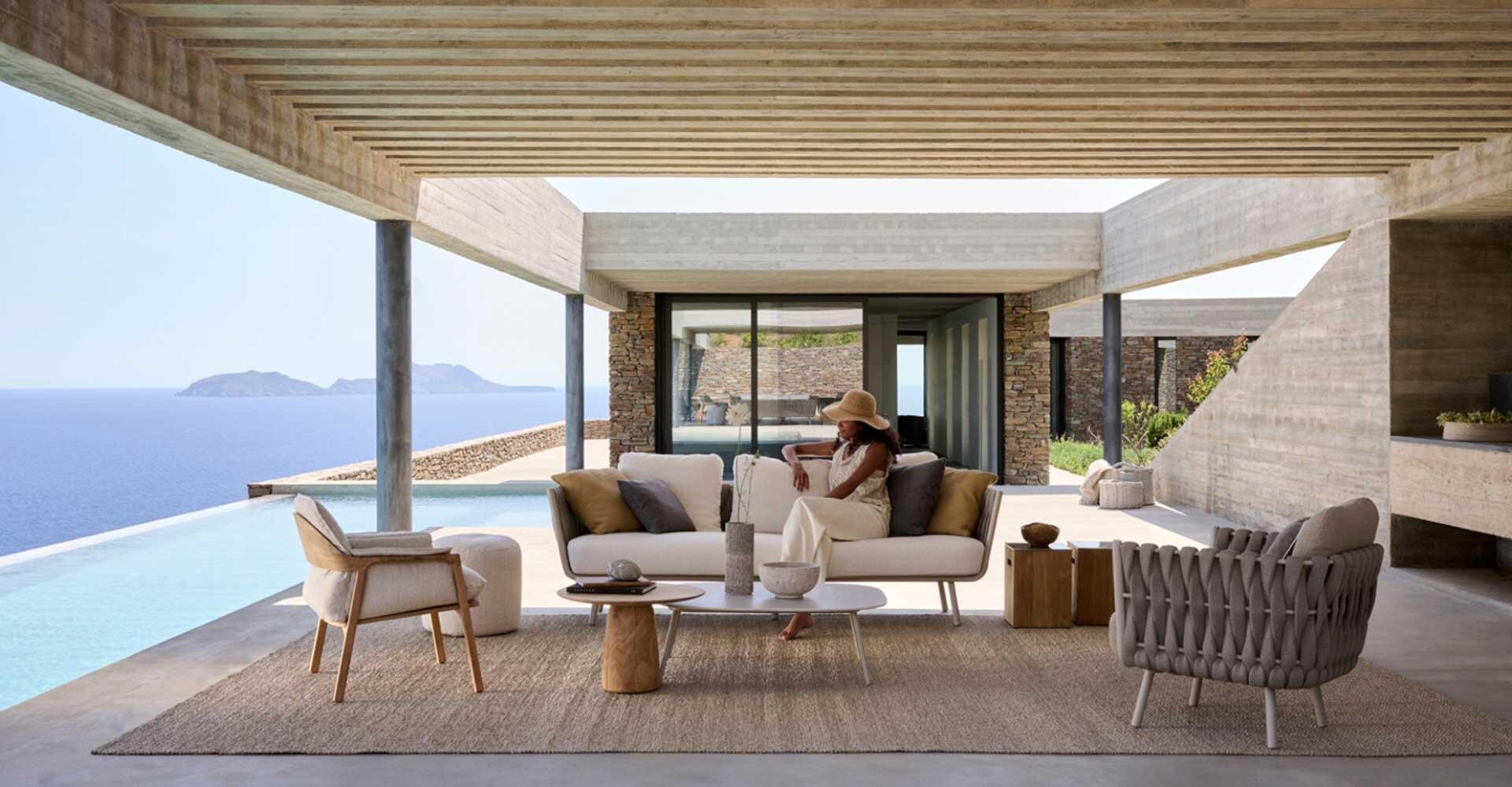 A woman in a sun hat sits on a modern outdoor sofa in a luxurious open terrace area overlooking an infinity pool and the ocean, with picturesque islands in the distance.