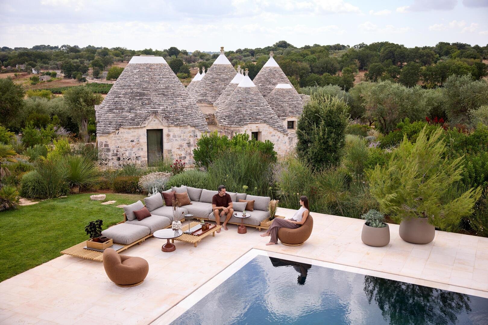 Two people relax on modern outdoor furniture next to a pool surrounded by lush greenery. In the background are traditional stone trulli houses with conical roofs, nestled in a rural landscape under a partly cloudy sky.