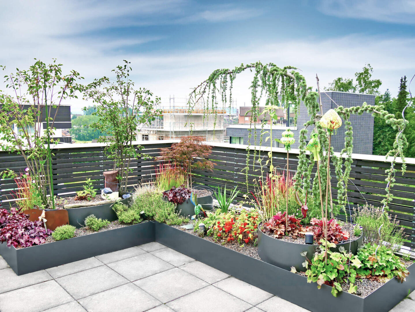 A modern rooftop garden with various green and leafy plants in raised beds, surrounded by a dark wooden fence, with buildings and a cloudy sky in the background.