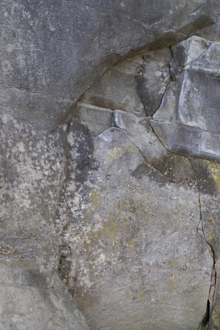 Nahaufnahme einer verwitterten Felsoberfläche mit sichtbaren Rissen, gezackten Kanten und Flecken von gelben und weißen Flechten. Der Fels zeigt natürliche Muster und Strukturen, die durch Erosion entstanden sind.