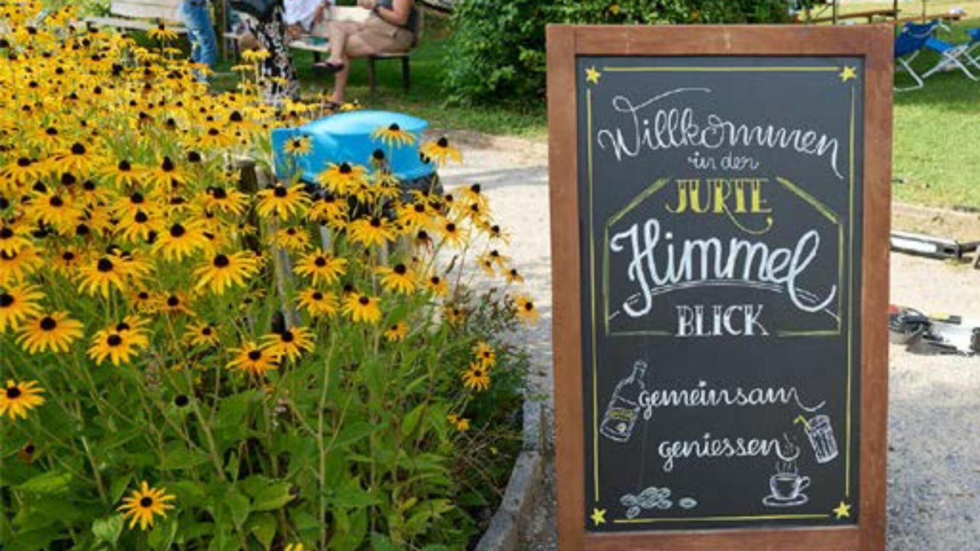 Ein Kreideschild in deutscher Sprache steht auf einem Kiesweg neben blühenden gelben Blumen, während im Hintergrund an einem sonnigen Tag Menschen an Tischen sitzen. Das Schild begrüßt die Besucher und ist mit handgezeichneten Verzierungen versehen.
