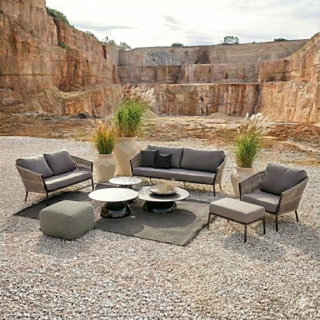 Lounge-Bereich im Freien auf Kies in einem Steinbruch mit grauen gepolsterten Sofas, Sesseln, einem Pouf, zwei niedrigen Tischen und großen Pflanzgefäßen mit hohen Gräsern, die alle auf einem gewebten Teppich angeordnet sind.