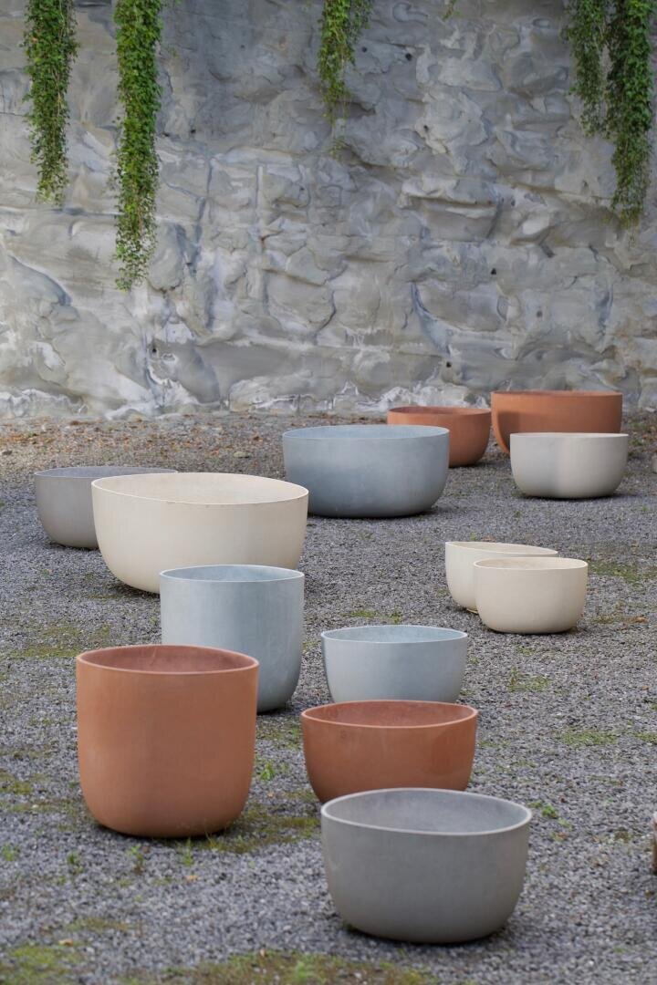 Large, empty plant pots in shades of terracotta, beige, and gray are arranged on gravel in front of a stone wall with green vines hanging down.