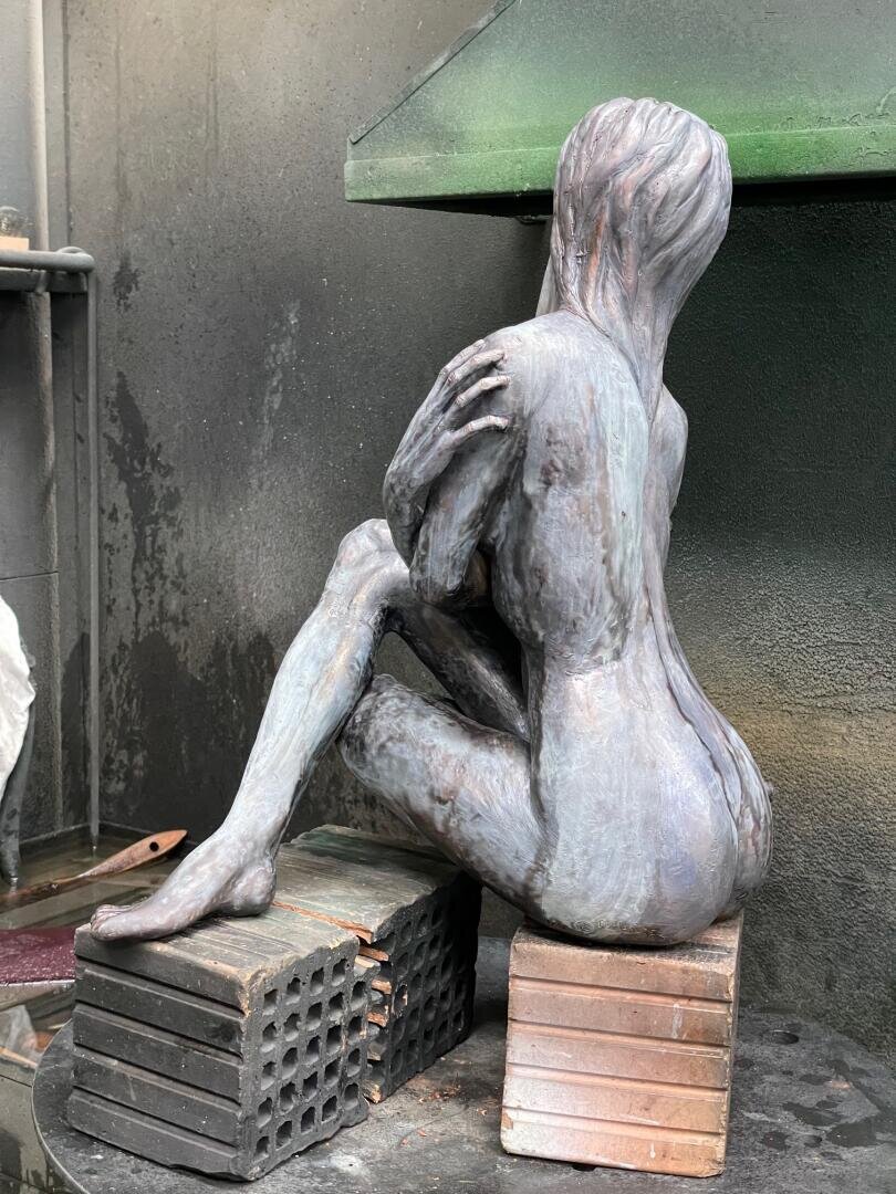 Bronzeskulptur einer sitzenden, nackten menschlichen Figur mit langem Haar, von hinten gesehen. Die Figur sitzt auf gestapelten Metall- und Ziegelblöcken in einer industriellen Werkstattumgebung.