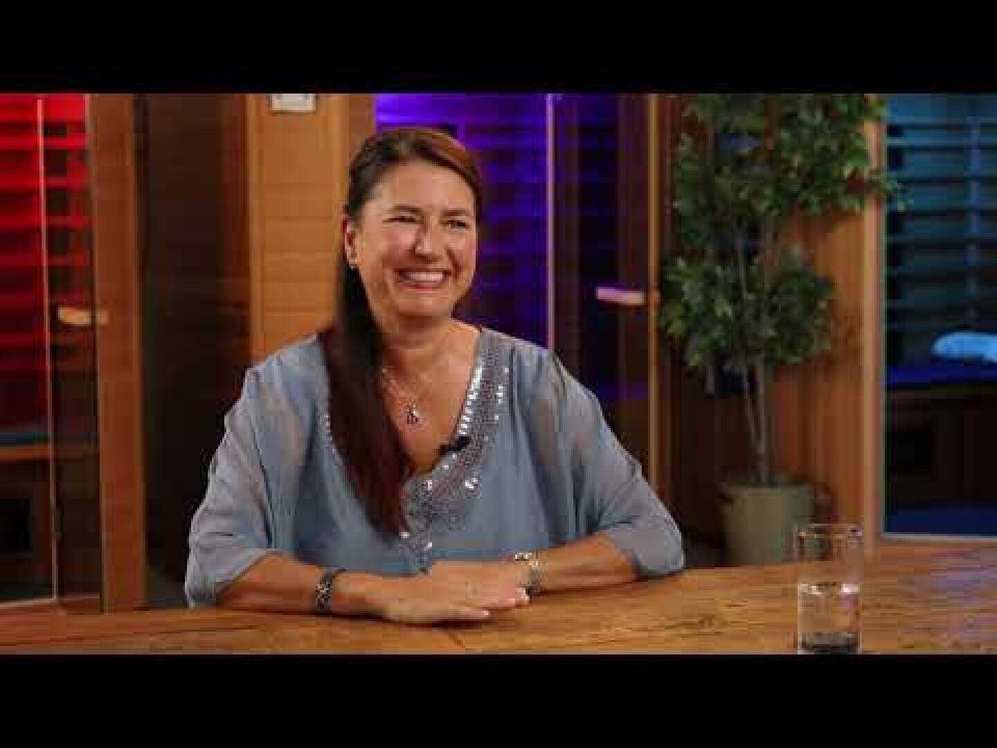 Eine Frau mit langen braunen Haaren und einer hellblauen Bluse sitzt lächelnd an einem Holztisch im Inneren des Raumes. Auf dem Tisch steht ein Glas Wasser und im Hintergrund befinden sich farbige, verschwommene Lichter und eine Topfpflanze.