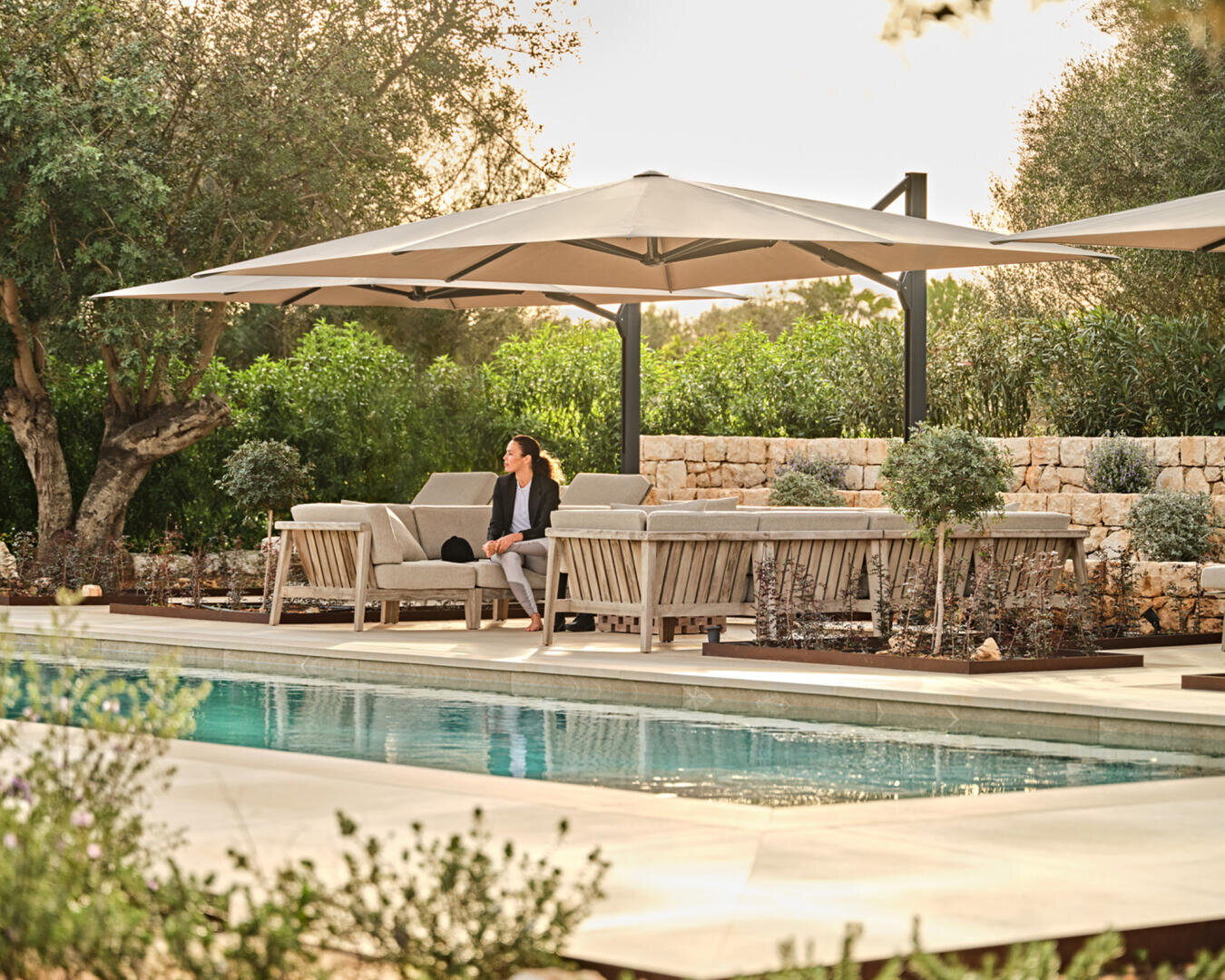 Eine Frau sitzt auf einem Sofa mit einem Laptop an einem Swimmingpool, unter großen Sonnenschirmen in einem Landschaftsgarten mit Bäumen, Sträuchern und einer Steinmauer im Hintergrund.