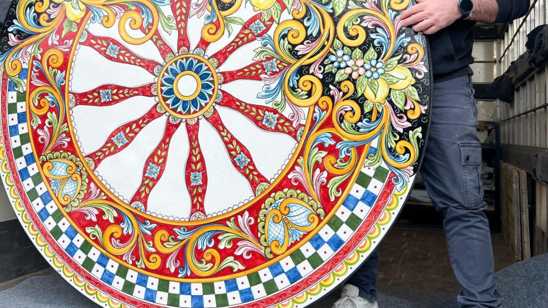 Eine Person hält ein großes, rundes, verziertes Objekt mit leuchtenden, verschlungenen Mustern in Rot, Gelb, Blau, Grün und Schwarz, die an traditionelle sizilianische Karrenkunst erinnern. Nur ein Teil der Person ist sichtbar.