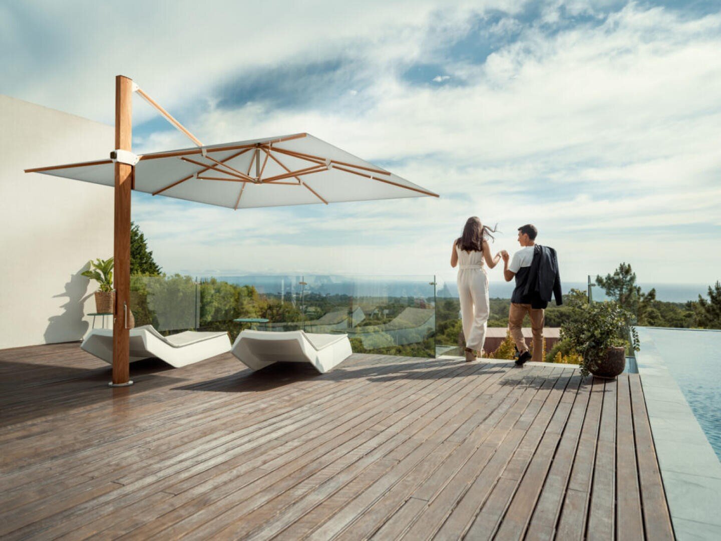 Ein händchenhaltendes Paar geht auf ein Glasgeländer auf einer Holzterrasse mit Liegestühlen, einem großen Sonnenschirm und einem Infinity-Pool zu und blickt auf eine malerische Landschaft unter einem teilweise bewölkten Himmel.