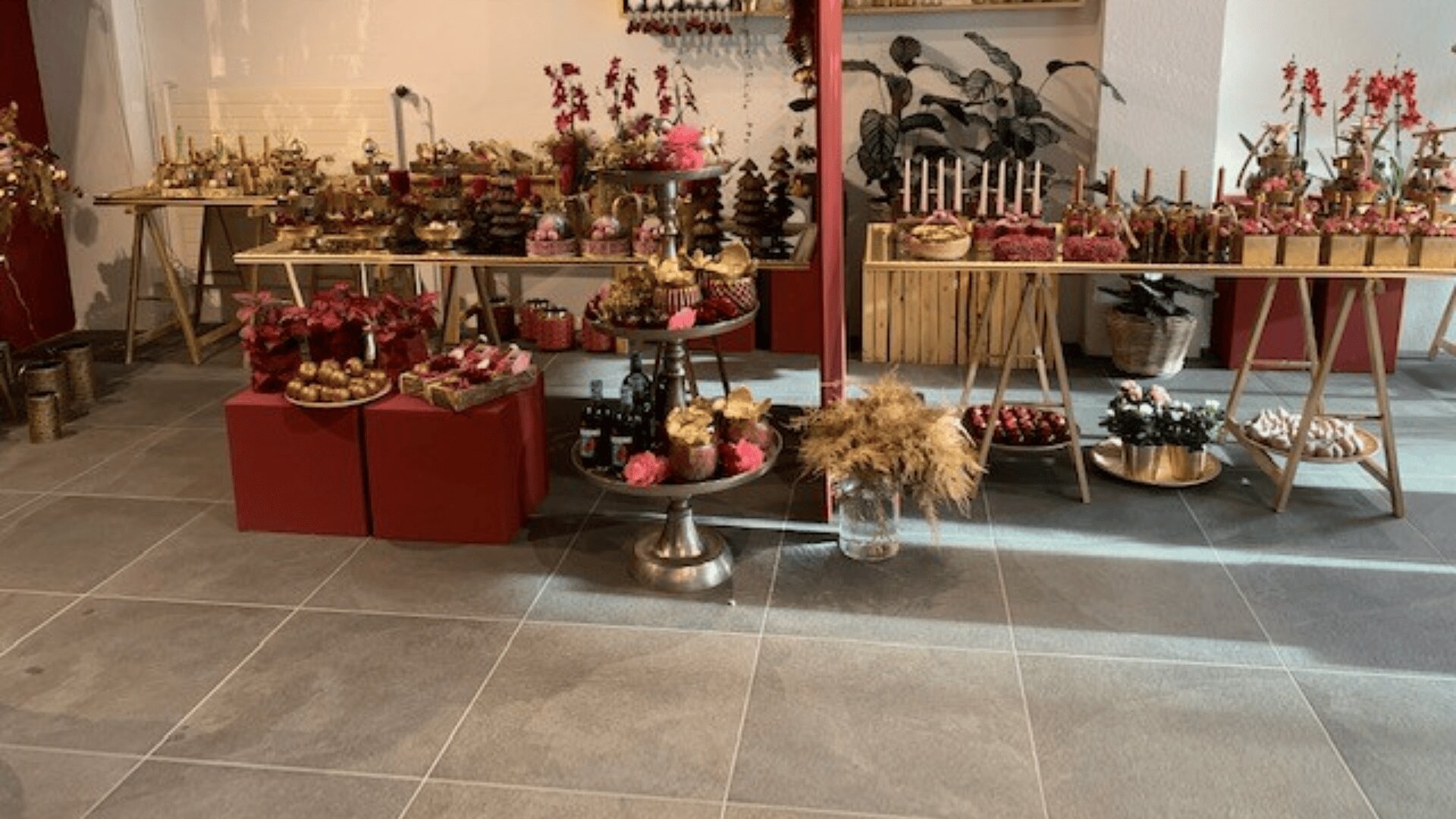Ein Desserttisch mit verschiedenen Süßigkeiten, Kuchen und dekorativen Blumen, die auf Ständern und Tischen angeordnet sind. Die Einrichtung besteht aus goldenen Akzenten und rosa Blumenarrangements vor einem gefliesten Boden und einer weißen Wand.