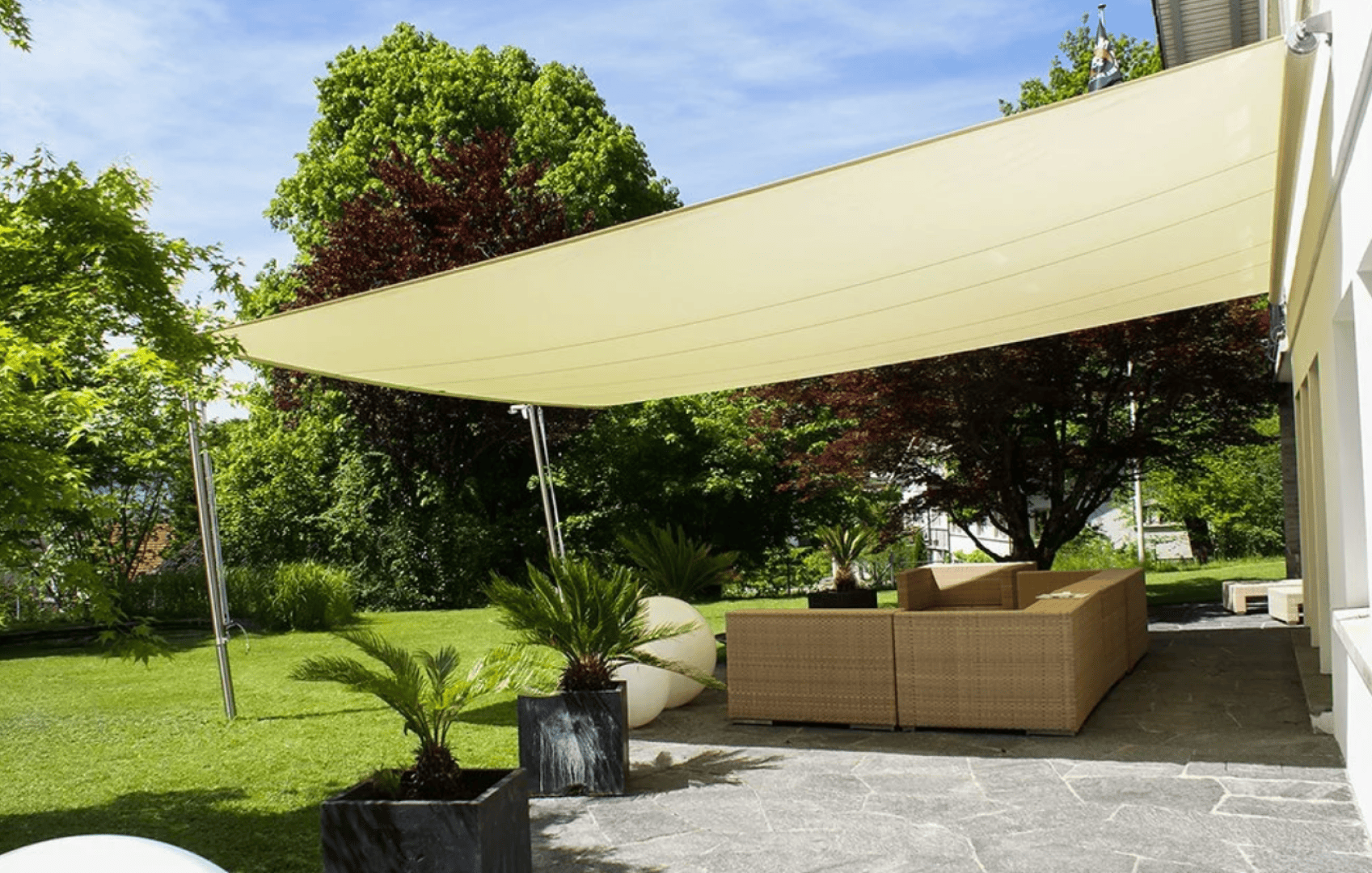 A modern outdoor patio with a beige shade sail overhead, wicker furniture with cushions, potted plants, and a garden with green trees and grass under a clear blue sky.