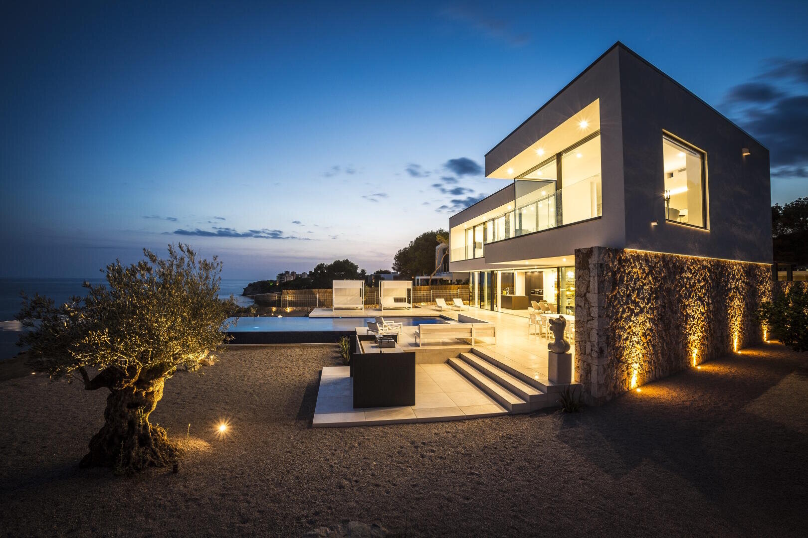 Modernes Haus mit großen Fenstern und Außenterrasse, beleuchtet in der Abenddämmerung, mit Blick auf ein ruhiges Meer. Die Szene zeichnet sich durch eine minimalistische Landschaftsgestaltung mit einem Baum und dekorativen Lichtern aus, die die Architektur akzentuieren.