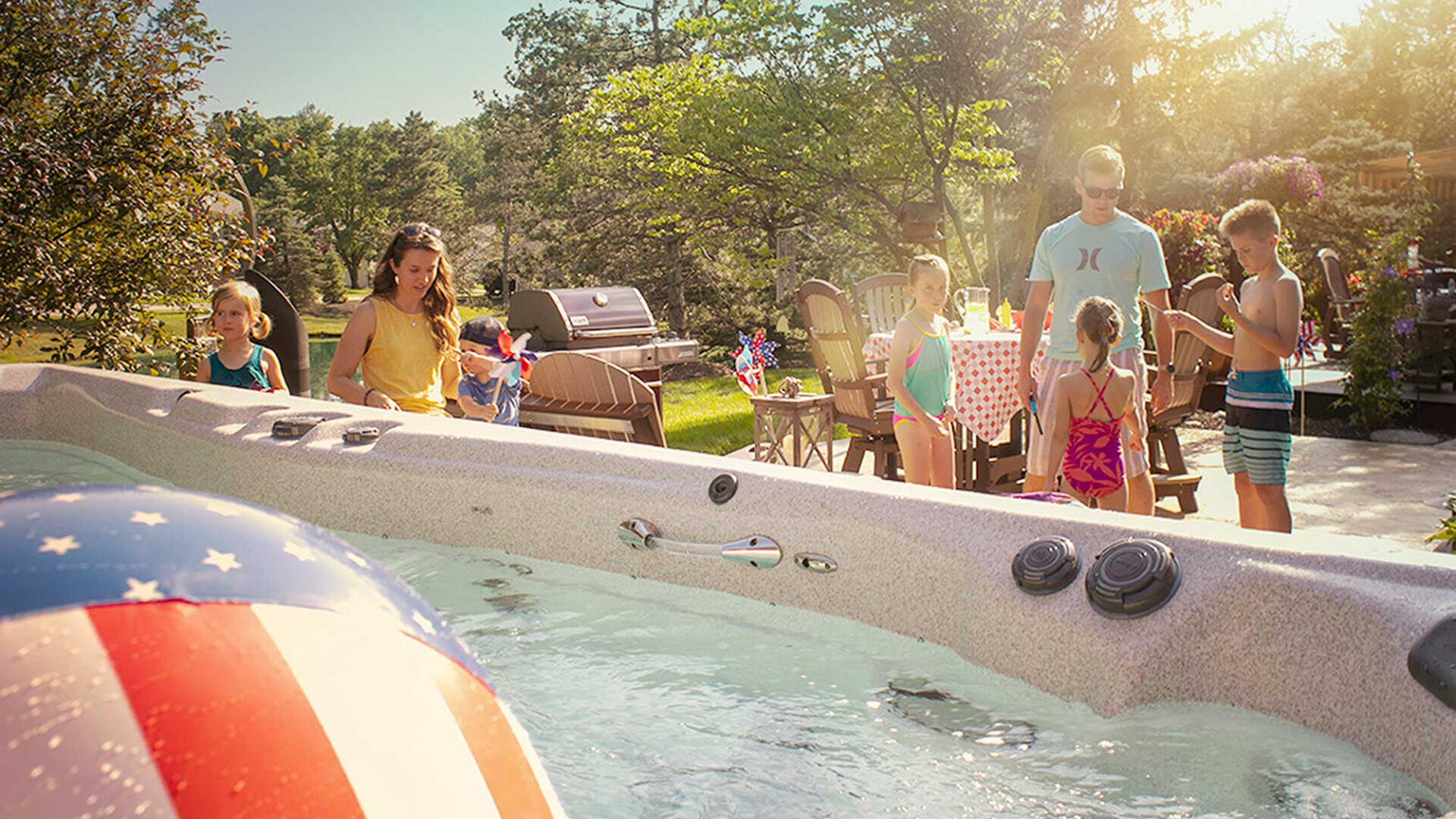 Eine Familie versammelt sich an einem sonnigen Tag im Freien an einem Whirlpool, wobei die Kinder am Wasser spielen und die Erwachsenen an einem für ein Picknick gedeckten Tisch stehen. Ein aufblasbarer Ball mit einem amerikanischen Flaggenmotiv schwimmt in der Wanne.