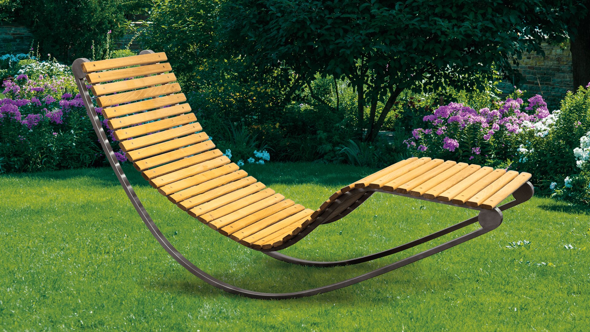 Ein hölzerner Schaukelstuhl mit einem geschwungenen Metallrahmen steht auf einem grünen Rasen, umgeben von blühenden Blumen und üppigen Bäumen in einem Garten.