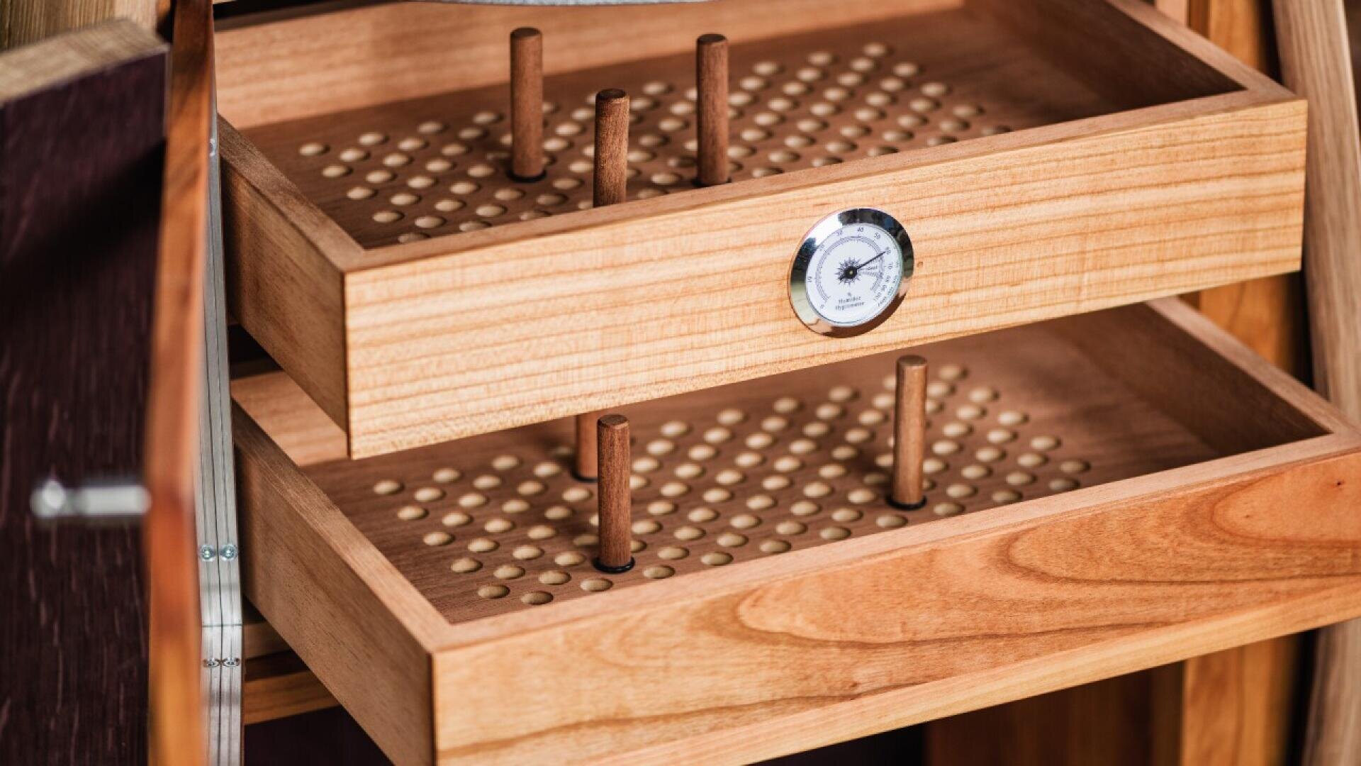 Open wooden humidor drawers with small holes, vertical wooden pegs, and a built-in analog hygrometer in the center of the top drawer, used for storing cigars.