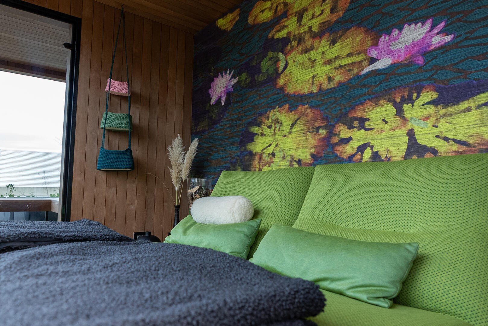 Ein gemütliches Schlafzimmer mit einem grünen Bett, grünen Kissen und einem weißen Nackenkissen. An der Holzwand ist eine bunte Wandmalerei mit Seerosen zu sehen, und auf der linken Seite befinden sich ein Fenster und hängende Körbe. Eine weiche graue Decke bedeckt das Bett.
