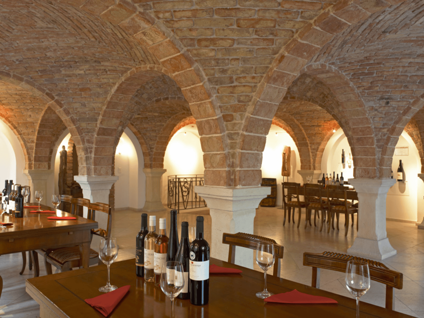 A wine cellar with arched brick ceilings, wooden tables set with wine bottles, glasses, and red napkins. Warm lighting highlights the stone columns and cozy, rustic atmosphere.