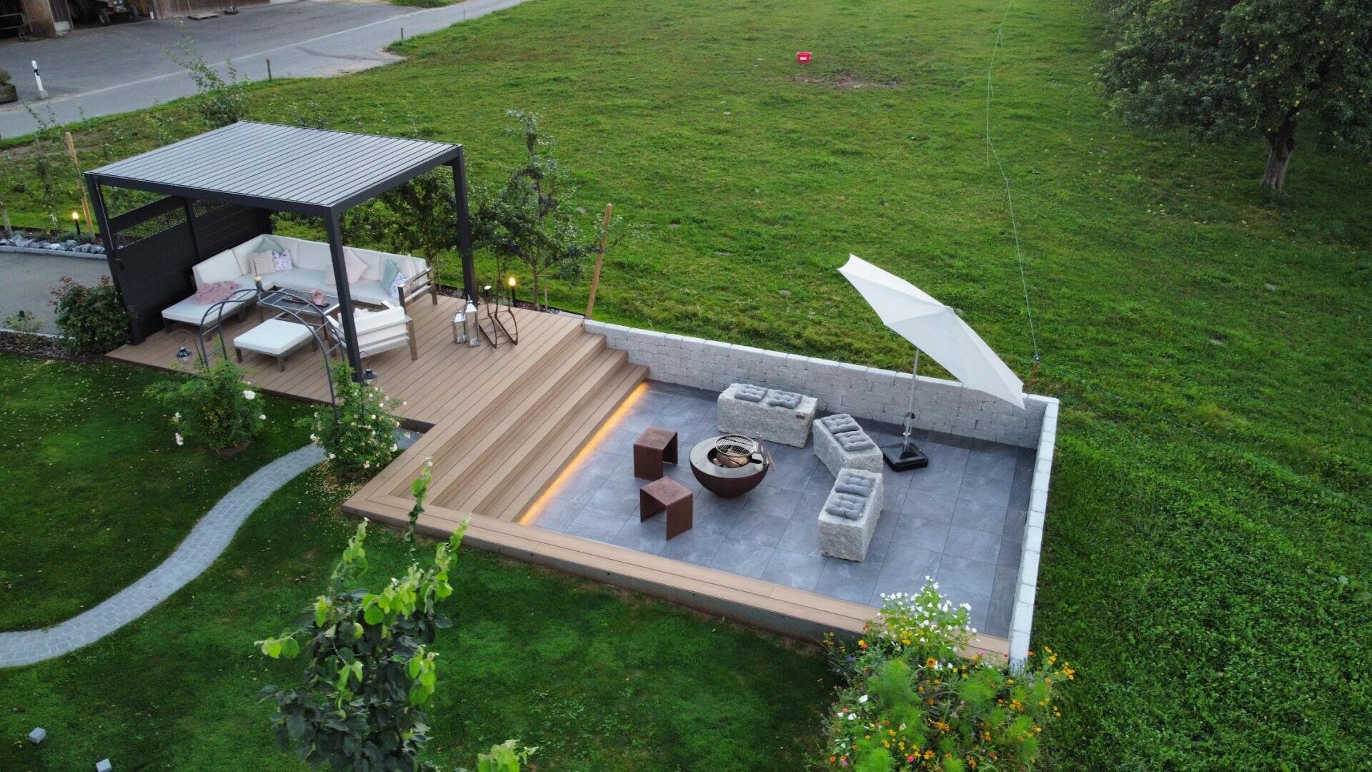 Ein moderner Hinterhof mit einem überdachten Sitzbereich auf einer Holzterrasse, Stufen, die hinunter zu einer versenkten Feuerstelle führen, Lounge mit grauen Sofas, Sesseln und einem großen weißen Sonnenschirm, umgeben von grünem Gras.