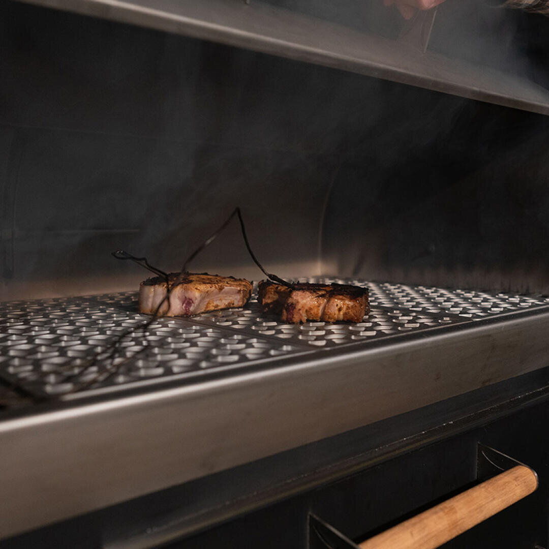 Two seasoned pieces of meat are cooking on a metal grill rack inside a smoker or grill, with smoke rising around them and a meat thermometer probe inserted in one of the pieces.