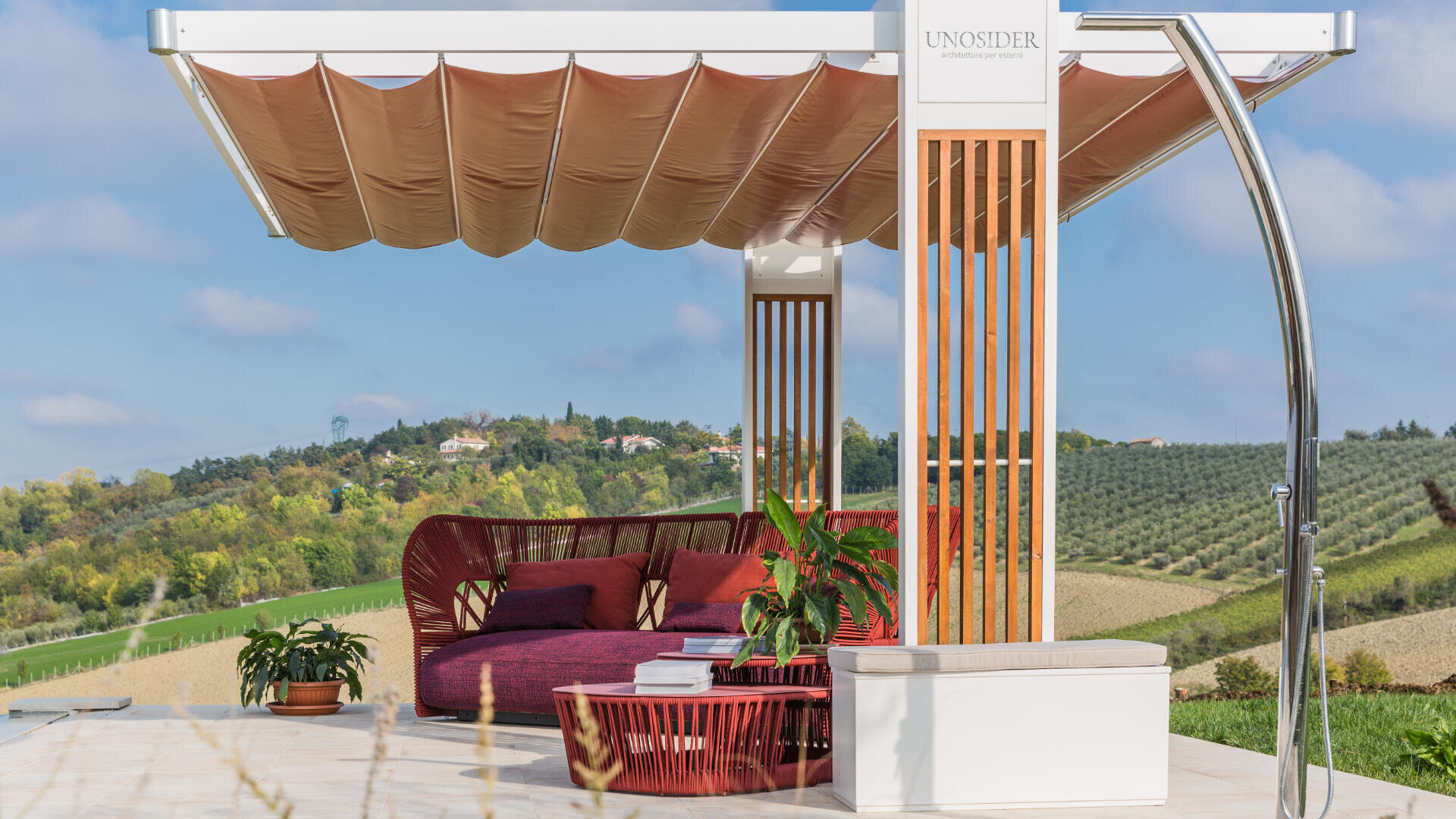 Eine moderne Outdoor-Lounge mit roten Sitzmöbeln, Topfpflanzen und einer hölzernen Pergola auf einer Terrasse mit Blick auf sanfte grüne Hügel und weit entfernte Häuser unter blauem Himmel.