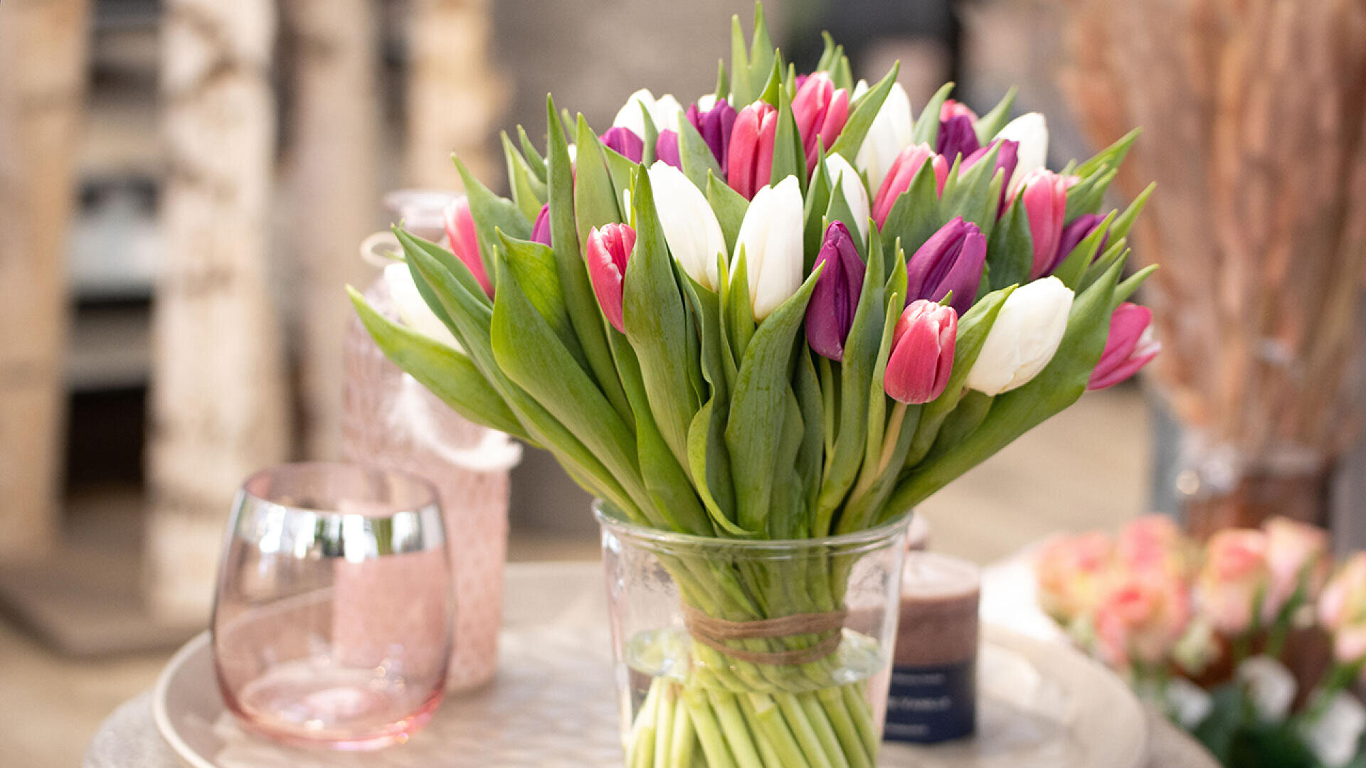 A clear glass vase filled with a bouquet of pink, white and purple tulips stands on a round table. A pink-tinted glass and blurred flowers stand nearby in the background.