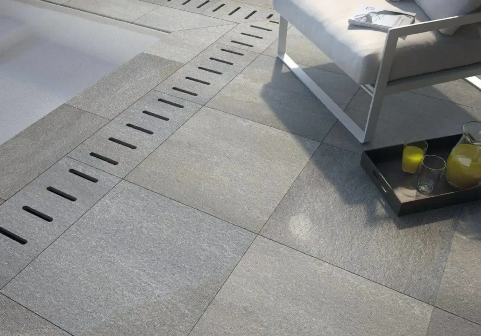 Moderne Außenterrasse mit großen grauen Steinfliesen, einem geradlinigen Abflussgitter aus Metall, einem weißen gepolsterten Stuhl und einem Tablett mit einem Glaskrug mit Saft und zwei Gläsern.