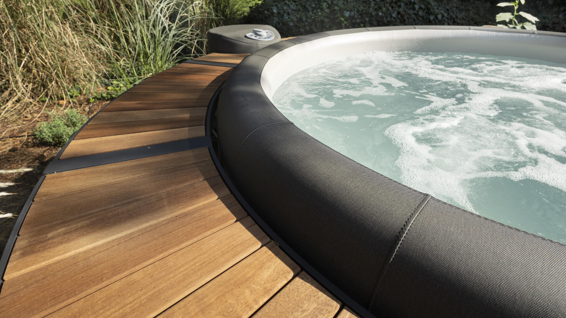Close-up of a bubbling outdoor hot tub with black trim, surrounded by a curved wooden deck and green plants in the background.