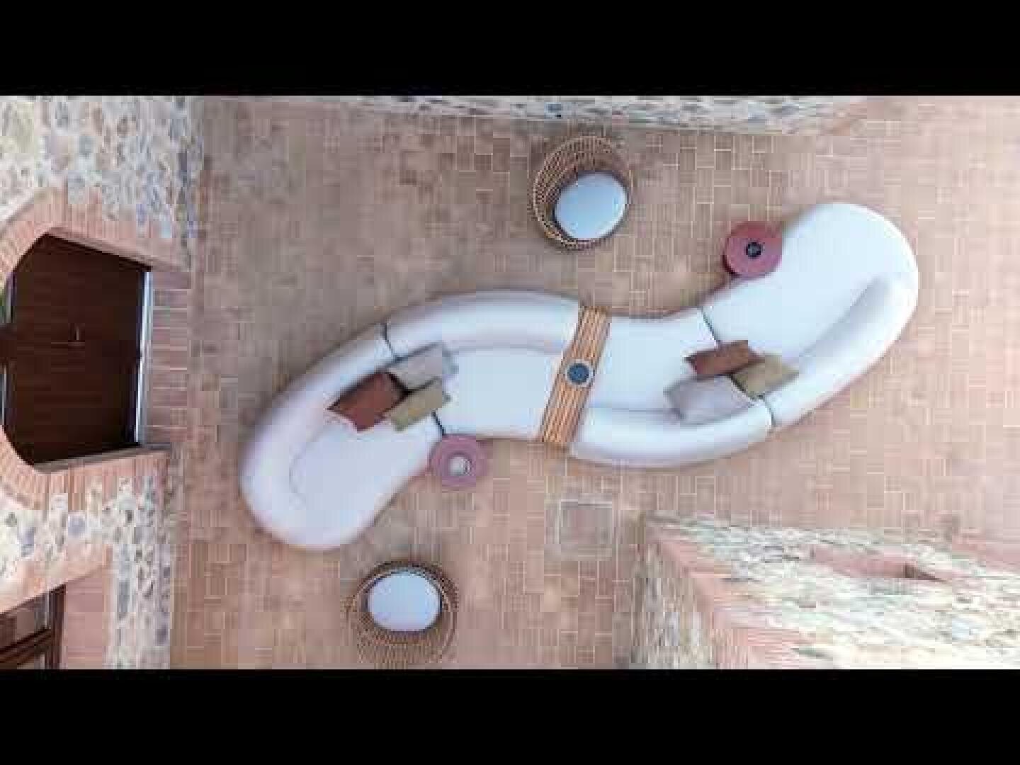 Curved white outdoor sofas with colorful cushions form an S-shape on a brick terrace. Nearby are three round tables with two wicker chairs, and stone walls surround the cozy seating area.