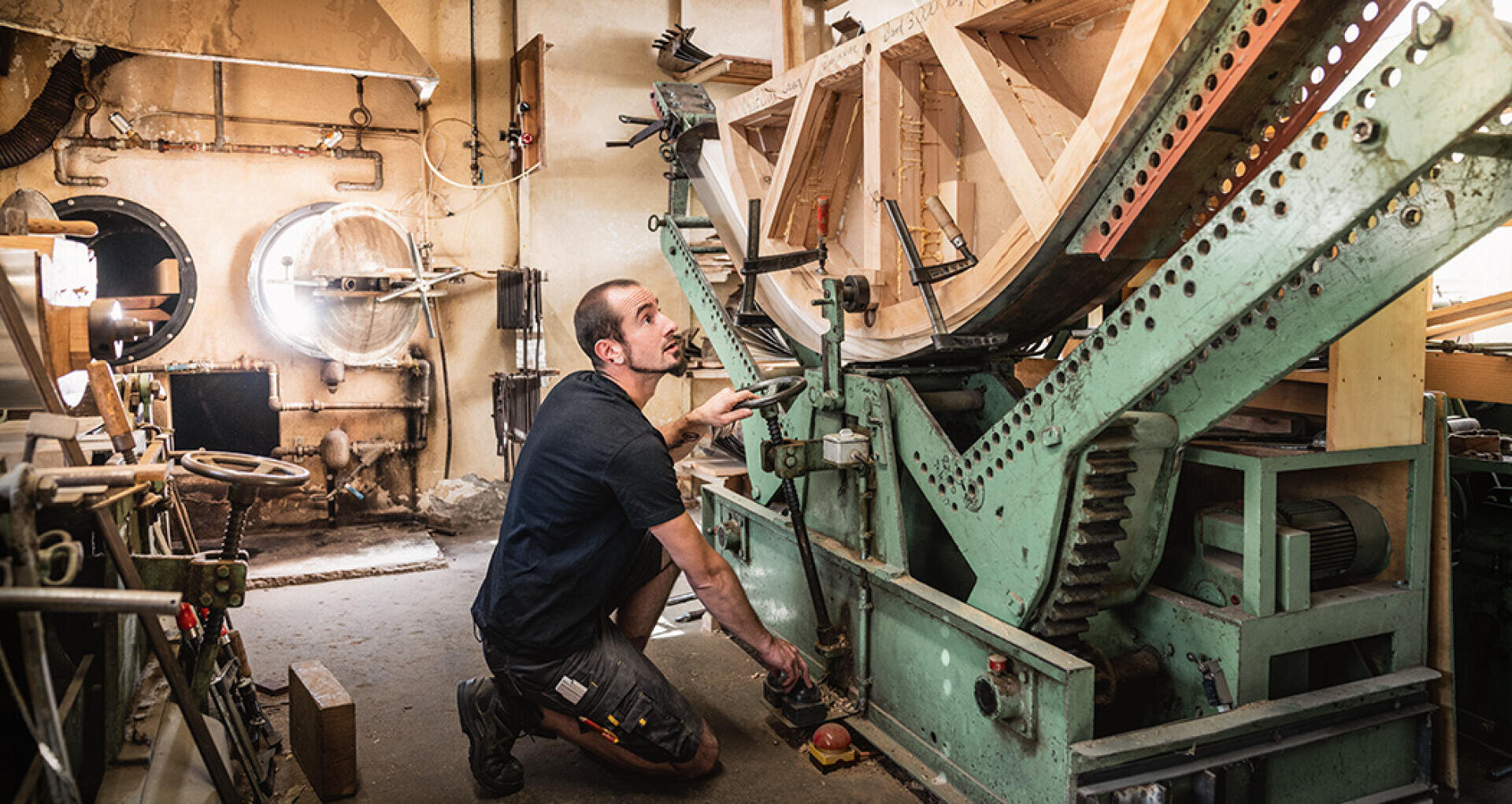 A man kneels next to a large, green industrial machine in a workshop and adjusts its controls. Wooden parts and mechanical components surround him, and sunlight streams in through a round window.
